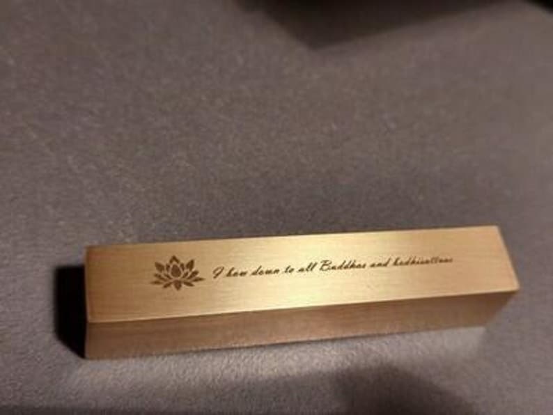 May include: A gold-colored metal bar with a lotus flower design and the text "I bow down to all Buddhas and bodhisattvas".