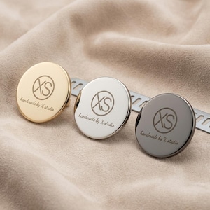 Può includere: Tre bottoni metallici circolari con finiture oro, argento e grigio scuro. Ogni bottone presenta un logo inciso con le lettere XS all'interno di un cerchio e il testo handmade by X studio sotto. Una striscia metallica è visibile dietro.