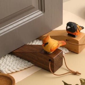 May include: Two wooden doorstops shaped like birds. One is a dark walnut wedge with a yellow bird featuring an orange beak. The other is a lighter wood wedge with a black and orange bird. Both have a small hole with a thin brown leather cord attached.