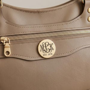 Könnte beinhalten: Eine taupefarbene Handtasche mit einem goldfarbenen Reißverschluss und einem runden, goldfarbenen Metall-Emblem in der Mitte der Vorderseite. Das Emblem ist mit dem geschwungenen Monogramm MLK und dem Text EST. 1978 graviert.