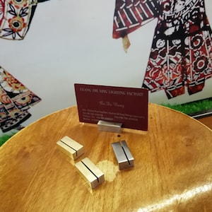 May include: Two gold and two silver metal business card holders on a wooden table. A business card with the text "Guang Zhi Xing Lighting Factory" is displayed in one of the holders.