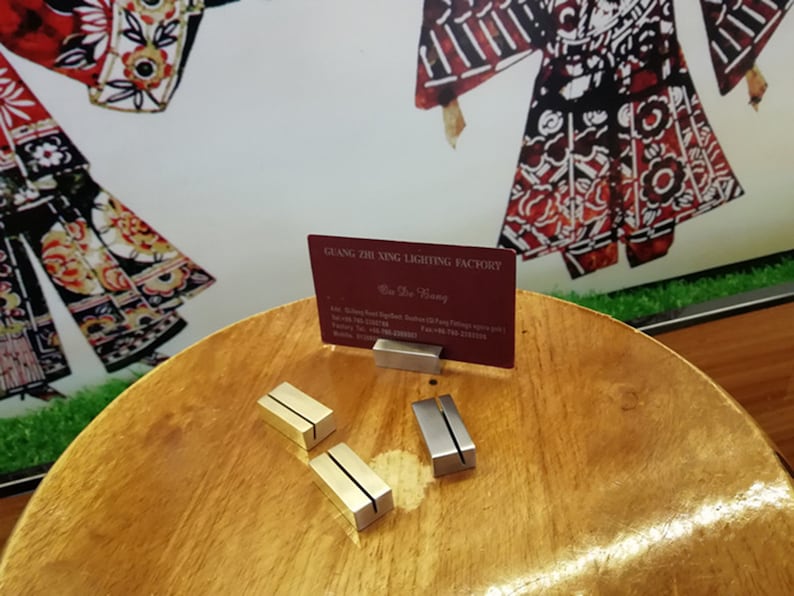 May include: Two gold and two silver metal business card holders on a wooden table. A business card with the text "Guang Zhi Xing Lighting Factory" is displayed in one of the holders.