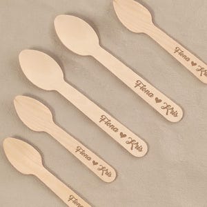 May include: Five wooden spoons with engraved text. The spoons are light brown and have a smooth finish. Each spoon has the name "Fiona" followed by a heart symbol and the name "Kris" engraved on the handle.