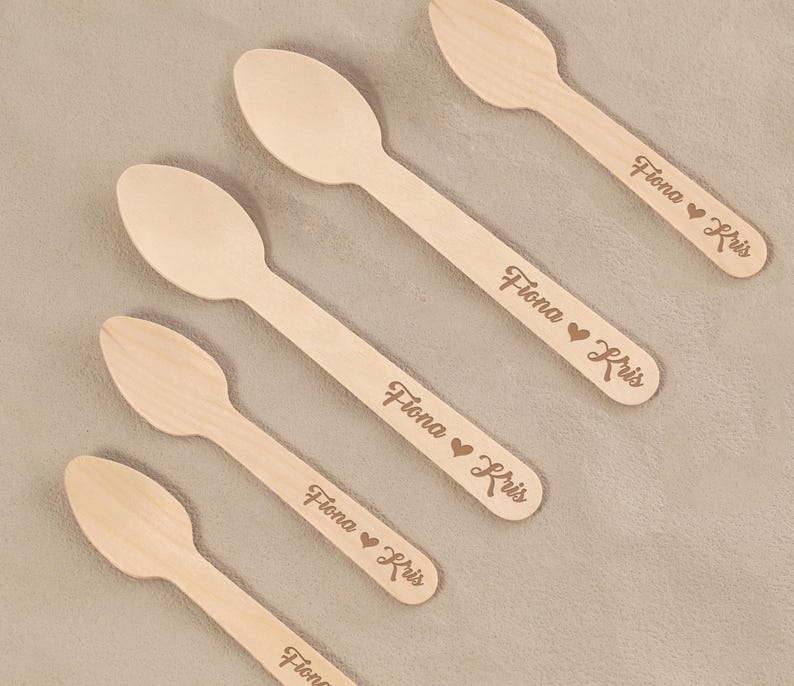 May include: Five wooden spoons with engraved text. The spoons are light brown and have a smooth finish. Each spoon has the name "Fiona" followed by a heart symbol and the name "Kris" engraved on the handle.