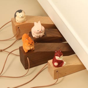 May include: Four wooden door stops featuring carved animal figures: a hedgehog, a rabbit, a squirrel, and a fox. Each wedge is made of light or dark wood, has a leather cord attached to the front, and is positioned to hold a white door open.