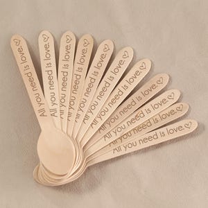 May include: A collection of wooden spoons, each engraved with the phrase "All you need is love" and a heart symbol. The spoons are arranged in a fan shape, showcasing their natural wood color and the delicate engraving.