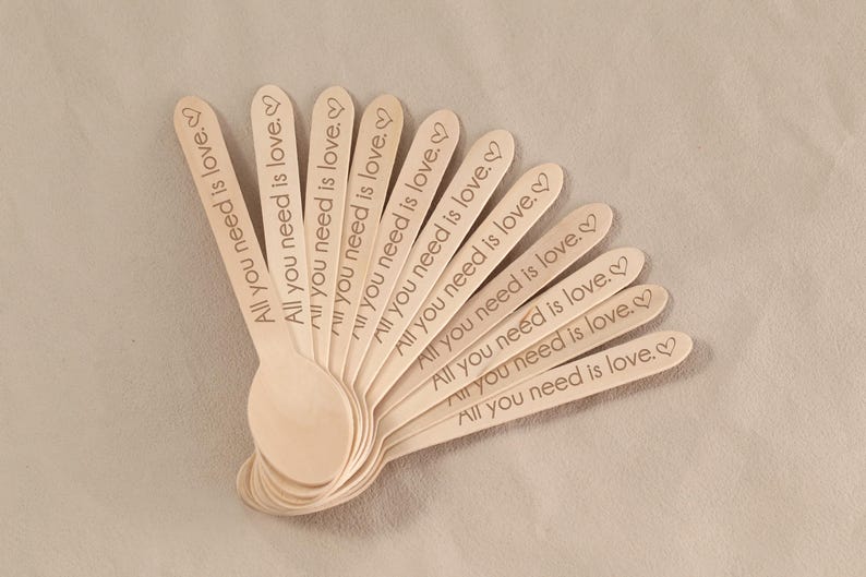 May include: A collection of wooden spoons, each engraved with the phrase "All you need is love" and a heart symbol. The spoons are arranged in a fan shape, showcasing their natural wood color and the delicate engraving.