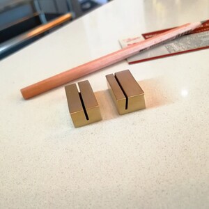 May include: Two gold metal card holders with a slot for a card. The holders are on a white surface.