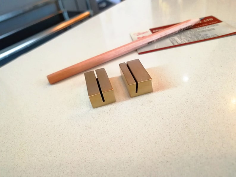 May include: Two gold metal card holders with a slot for a card. The holders are on a white surface.