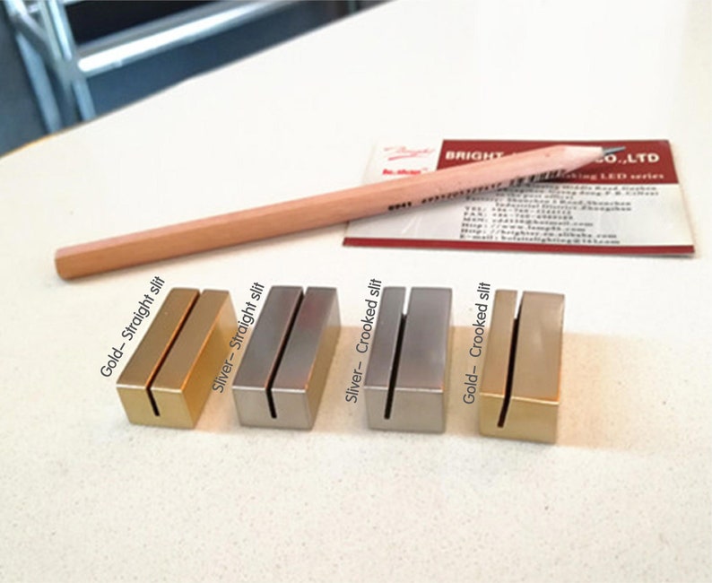 May include: Four metal table number holders, two gold and two silver. The holders are in two styles, straight and crooked. The text on the holders reads "Gold - Straight slit", "Silver - Straight slit", "Silver - Crooked slit", and "Gold - Crooked slit".