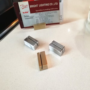 May include: Three table number holders, two are silver and one is gold. The holders are rectangular with a slot in the middle for a card.