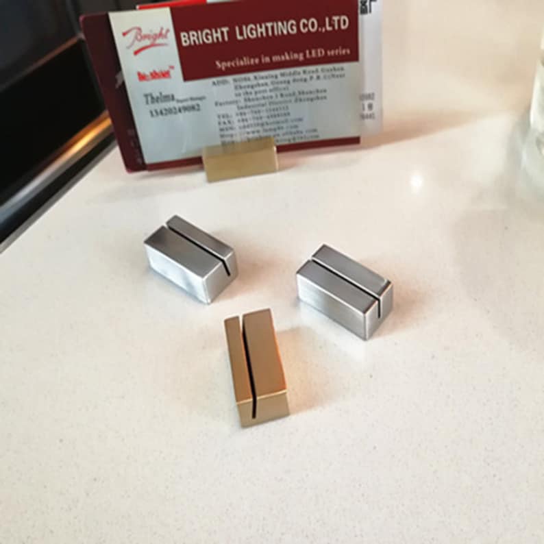 May include: Three table number holders, two are silver and one is gold. The holders are rectangular with a slot in the middle for a card.