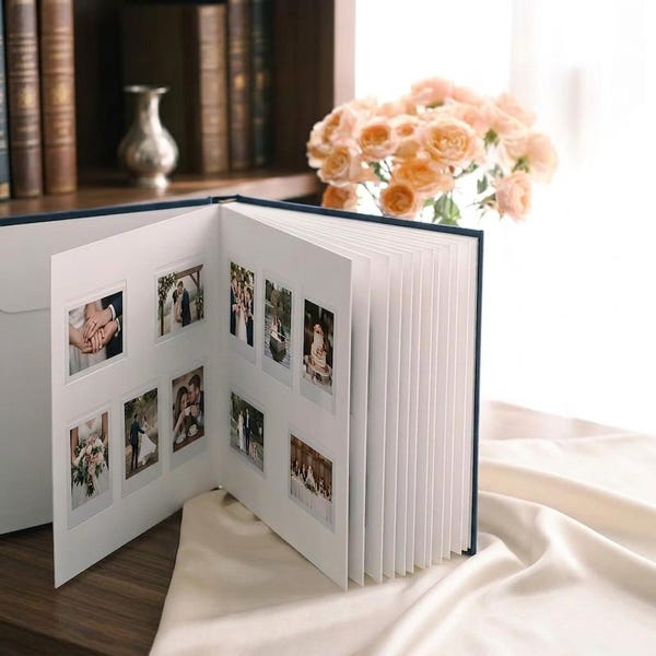 May include: A navy blue photo album with thick white pages, open to display multiple small square photographs of wedding scenes. The album features a hard cover and is positioned on a soft, draped cream-colored fabric surface.
