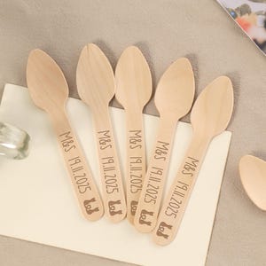 May include: A collection of wooden spoons, each engraved with "M&S 19.11.2025" and a small icon. The spoons are arranged on a white surface, with one spoon isolated. The spoons are light brown in color and appear to be disposable.