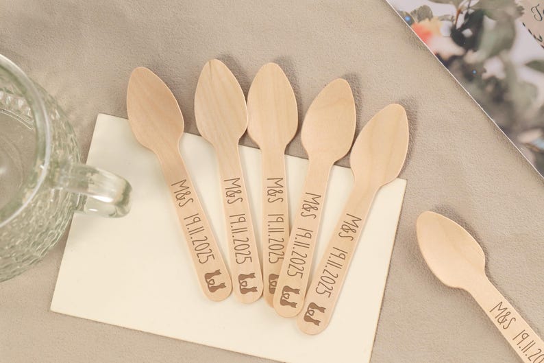 May include: A collection of wooden spoons, each engraved with "M&S 19.11.2025" and a small icon. The spoons are arranged on a white surface, with one spoon isolated. The spoons are light brown in color and appear to be disposable.