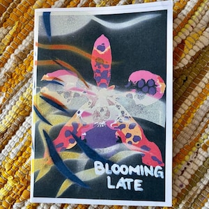 Può includere: Un biglietto d'auguri rettangolare con un'illustrazione stilizzata e colorata di un'orchidea su sfondo scuro. Il fiore presenta sfumature di rosa, viola e arancione con motivi a macchie. Il testo bianco recita BLOOMING LATE.