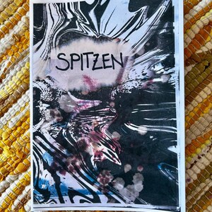 Può includere: Una stampa rettangolare con un design a inchiostro marmorizzato bianco e nero con vortici e schizzi astratti. La parola SPITZEN è stampata in lettere maiuscole nere al centro dell'immagine, su uno sfondo più chiaro e strutturato.
