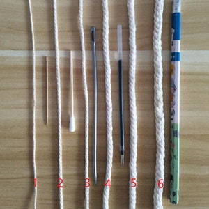 May include: Six white cords of varying thickness and textures, laid out horizontally on a wooden surface. A toothpick, cotton swab, metal needle, pen, and pencil are placed next to the cords for size comparison.