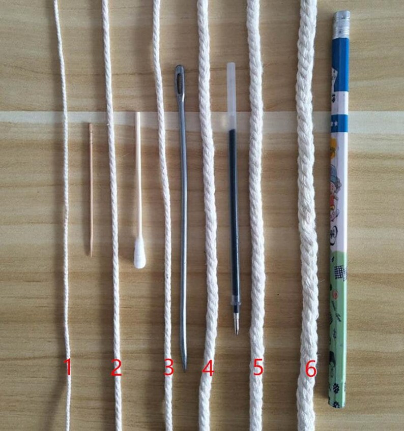 May include: Six white cords of varying thickness and textures, laid out horizontally on a wooden surface. A toothpick, cotton swab, metal needle, pen, and pencil are placed next to the cords for size comparison.
