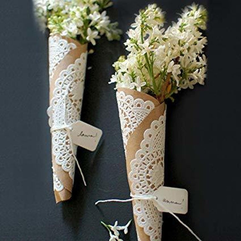 K&ouml;nnte beinhalten: Zwei Str&auml;u&szlig;e wei&szlig;er Blumen, die in braunes Papier und wei&szlig;e Spitzendeckchen gewickelt sind. Die Deckchen haben ein zartes Muster und sind mit Schnur gebunden. Jeder Strau&szlig; hat ein kleines wei&szlig;es Etikett mit handschriftlichem Text.