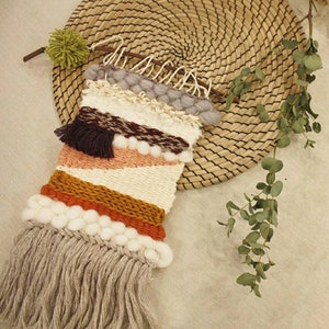 Pode incluir: Uma tape&ccedil;aria de parede tecida com uma borda de franjas. A tape&ccedil;aria apresenta um padr&atilde;o geom&eacute;trico em tons de marrom, laranja, rosa e branco. &Eacute; decorada com pompons brancos e uma borla marrom.