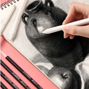 May include: A set of five black drawing pencils with the text "Artmark, Artist, C101" printed on each pencil. The pencils are arranged in a row on a pink surface. A drawing of a vase and fruit is in the background.