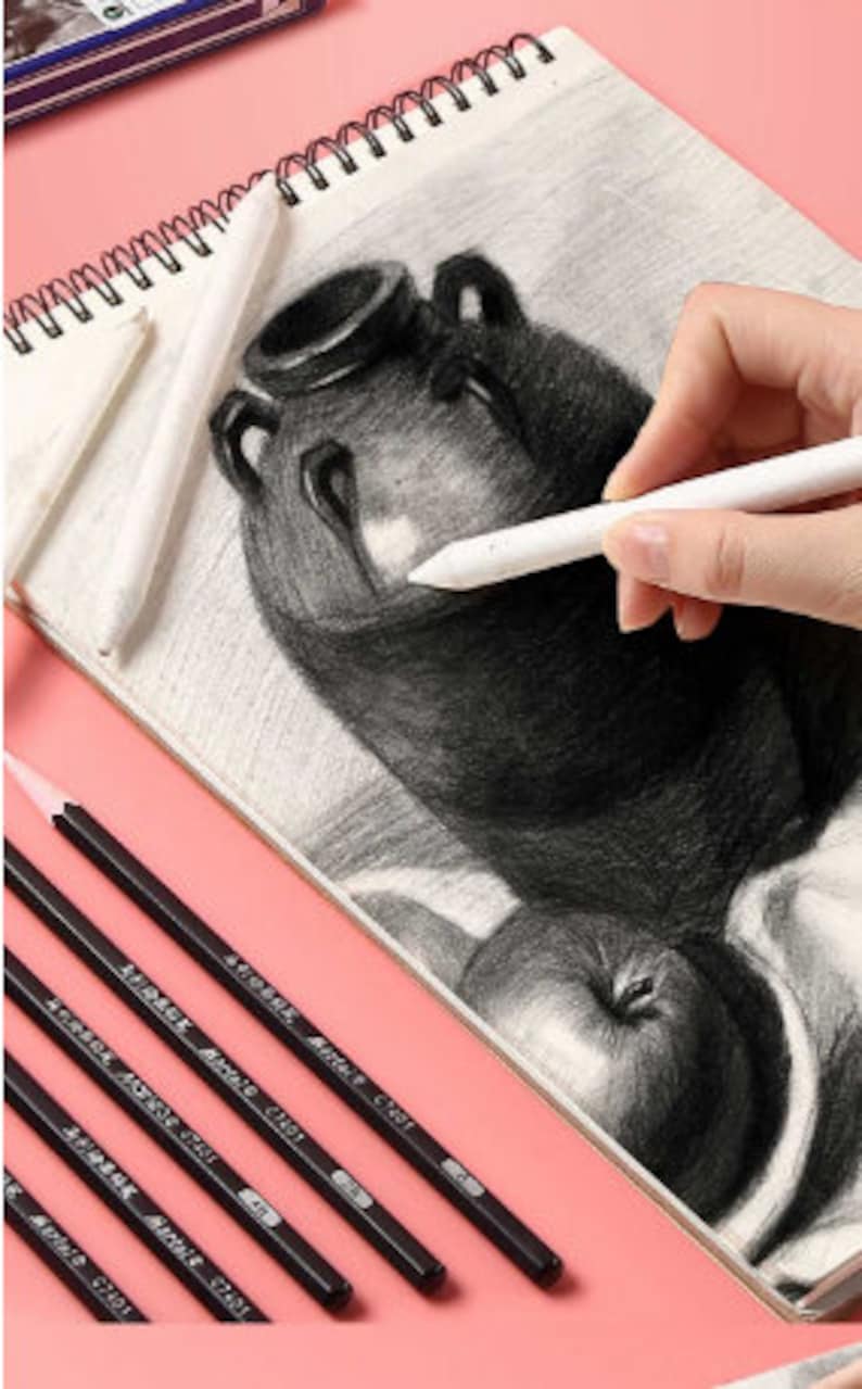 May include: A set of five black drawing pencils with the text "Artmark, Artist, C101" printed on each pencil. The pencils are arranged in a row on a pink surface. A drawing of a vase and fruit is in the background.