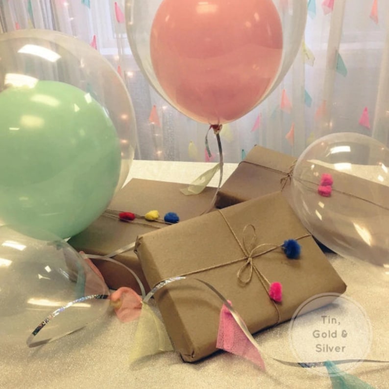 K&ouml;nnte beinhalten: Drei braune Geschenkboxen, die mit Schnur umwickelt und mit bunten Bommeln verziert sind. Die Boxen sind von durchsichtigen Luftballons umgeben, einer mit einem rosa Luftballon darin und der andere mit einem gr&uuml;nen Luftballon darin. Der Text "Tin, Gold & Silver" ist in der unteren rechten Ecke des Bildes sichtbar.