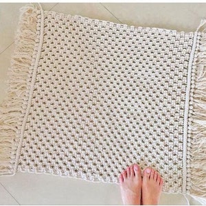 May include: A white macrame rug with fringe on all sides. The rug is made with a simple knot pattern and has a natural, bohemian style.