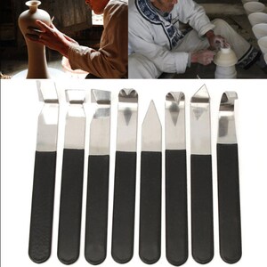 May include: Set of ten stainless steel pottery tools with black handles. The tools have various shapes and sizes for sculpting and trimming clay.