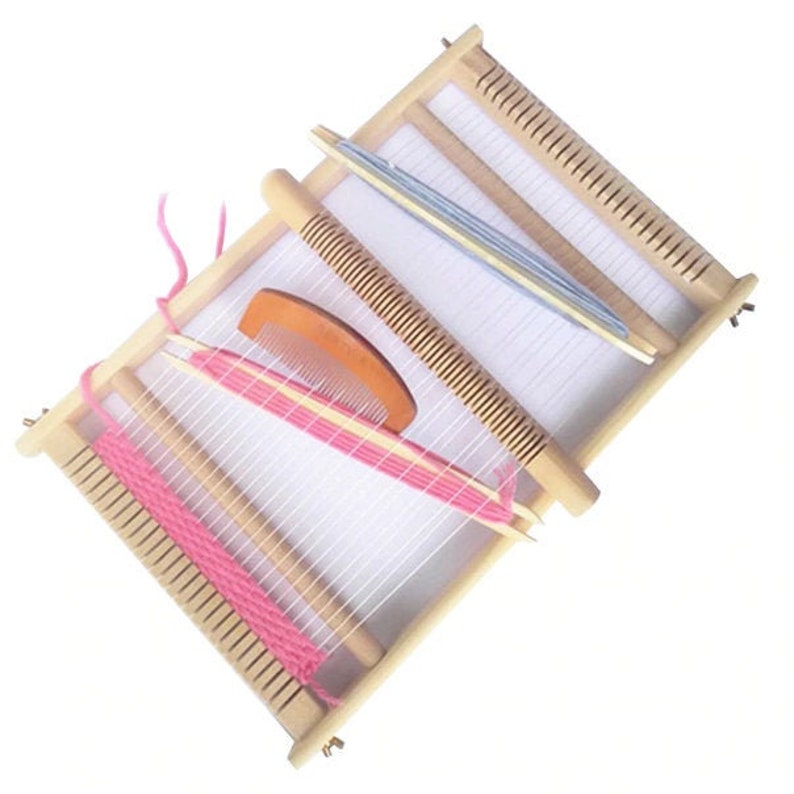 Pode incluir: Um tear de madeira com fio rosa e um pente para tecer. O tear tem um fundo branco e &eacute; feito de madeira natural.