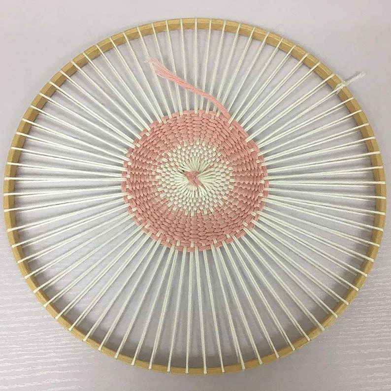 May include: A circular loom with white yarn wrapped around the spokes. A pink and white woven tapestry is partially completed in the centre of the loom.