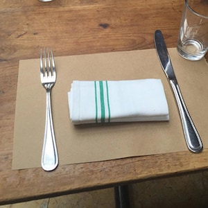 K&ouml;nnte beinhalten: Ein braunes Papiertischset mit einer wei&szlig;en Serviette mit gr&uuml;nen Streifen, einer silbernen Gabel und einem silbernen Messer auf einem Holztisch.