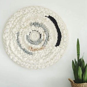 May include: A white, round wall hanging with a woven texture. The centre of the hanging features a circular design with shades of grey and pink. The outer edge of the hanging has a black stripe.