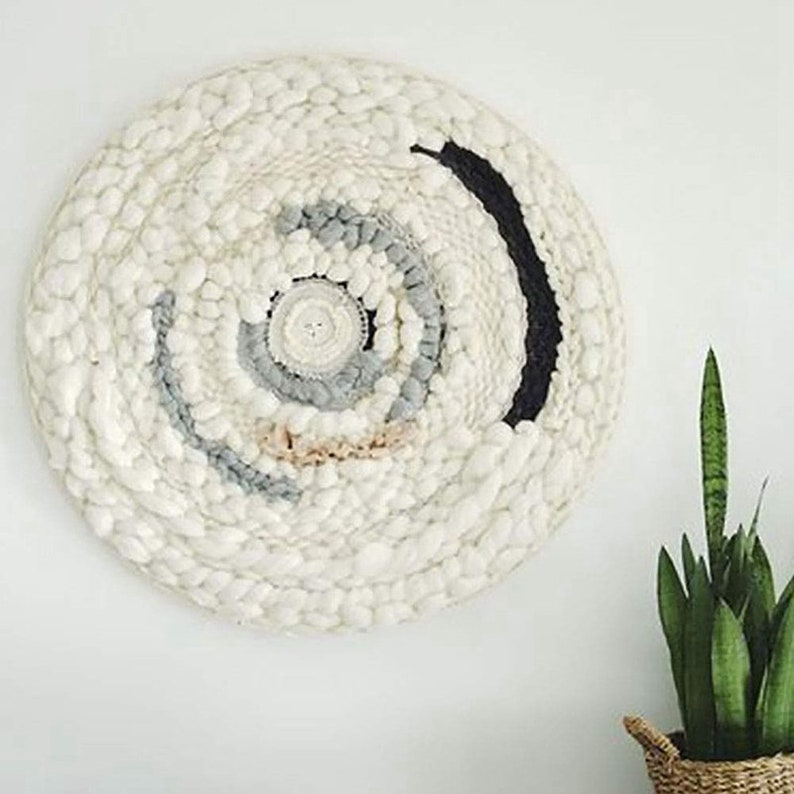 May include: A white, round wall hanging with a woven texture. The centre of the hanging features a circular design with shades of grey and pink. The outer edge of the hanging has a black stripe.