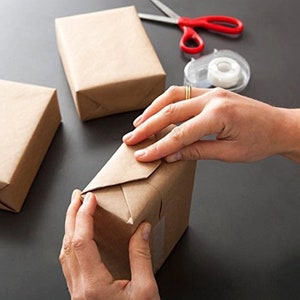 K&ouml;nnte beinhalten: Eine Person verpackt ein braunes Papierpaket mit Klebeband. Das Paket liegt auf einer schwarzen Oberfl&auml;che.