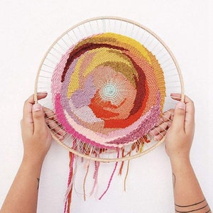 May include: A circular woven wall hanging with a colourful abstract design in shades of pink, orange, yellow, and brown. The hanging is made with yarn and is framed in a wooden hoop.