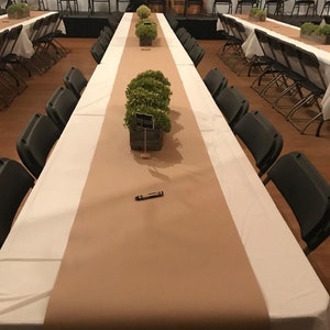 K&ouml;nnte beinhalten: Ein langer, brauner Tischl&auml;ufer mit gr&uuml;nen Pflanzen in einer Holzkiste auf einer wei&szlig;en Tischdecke. Der Tisch ist mit St&uuml;hlen und einem schwarzen Stift gedeckt.