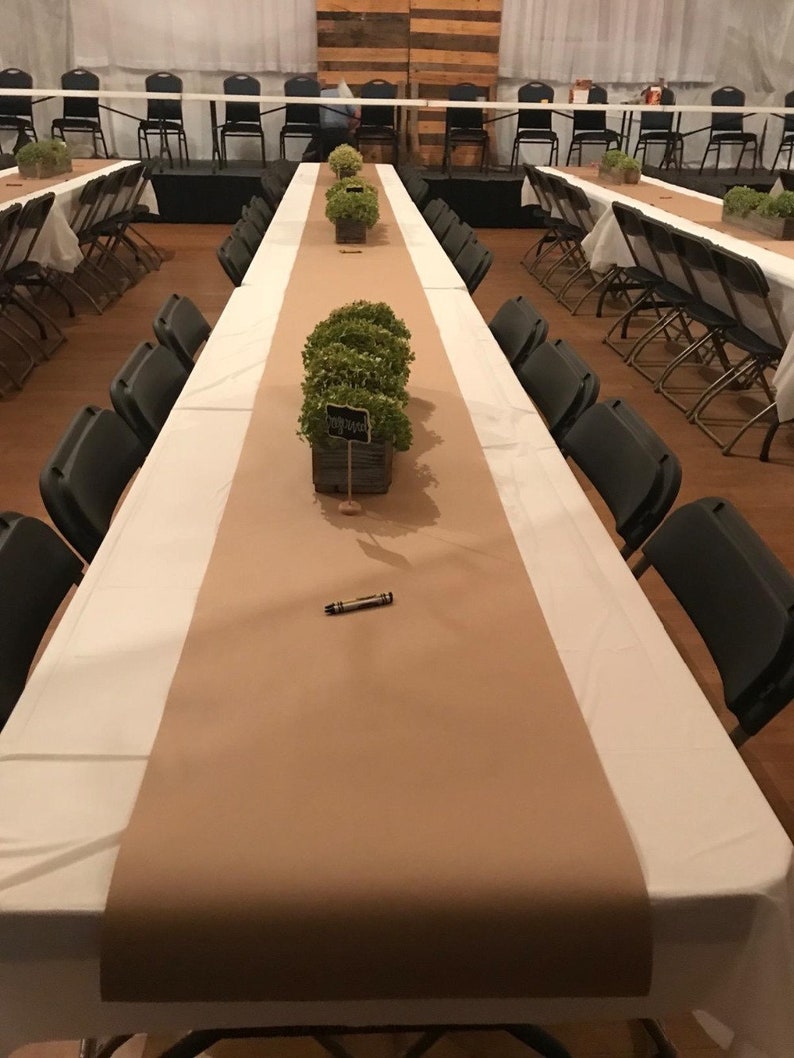 K&ouml;nnte beinhalten: Ein langer, brauner Tischl&auml;ufer mit gr&uuml;nen Pflanzen in einer Holzkiste auf einer wei&szlig;en Tischdecke. Der Tisch ist mit St&uuml;hlen und einem schwarzen Stift gedeckt.