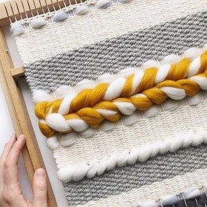 Pode incluir: Uma tape&ccedil;aria de parede tecida com um padr&atilde;o de listras cinzas e brancas. Uma tran&ccedil;a grossa de fio amarelo e branco &eacute; tecida no centro da tape&ccedil;aria.