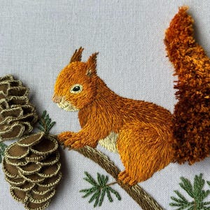 May include: A detailed embroidery piece on light gray fabric featuring a vibrant orange squirrel with a textured, fluffy tail perched on a brown branch. Two brown pinecones with green stitched needles are positioned to the left of the squirrel.