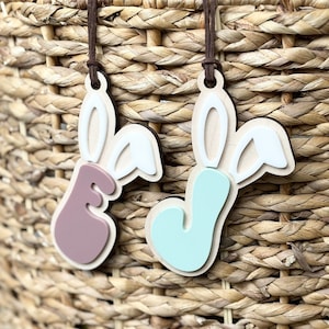 May include: Two wooden Easter bunny decorations with brown suede cords hang against a woven basket. One bunny has a lavender body, the other a light blue body. Both have white and natural wood accents.