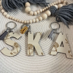 May include: Wooden keychains spelling out the word "SKA" in a light brown colour with a floral design in gold. Each letter has a small tassel attached to the top.