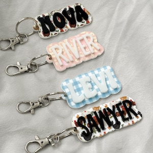 Op de afbeelding: Vier gepersonaliseerde sleutelhangers met de namen "NOVA", "RIVER", "LEVI" en "SAWYER". "NOVA" en "SAWYER" hebben een kippen- en bloemenpatroon. "RIVER" heeft een roze luipaardprint en "LEVI" een blauw ruitpatroon. Zilverkleurige karabijnsluitingen.