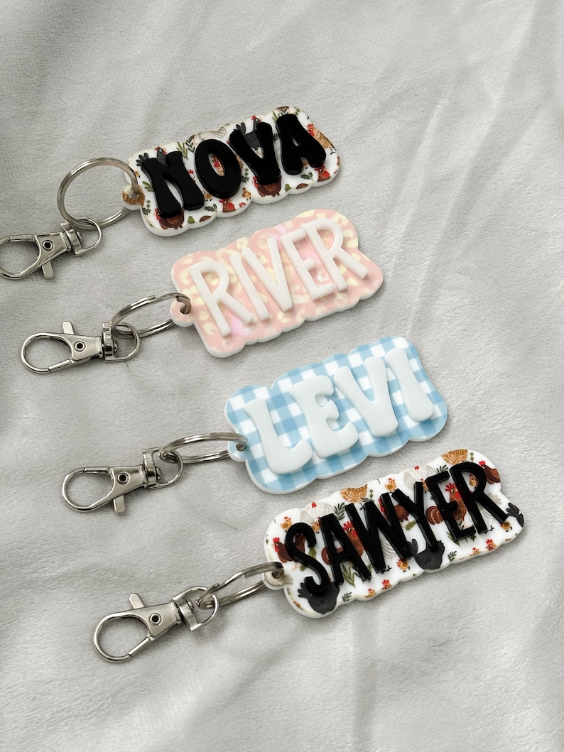 Op de afbeelding: Vier gepersonaliseerde sleutelhangers met de namen "NOVA", "RIVER", "LEVI" en "SAWYER". "NOVA" en "SAWYER" hebben een kippen- en bloemenpatroon. "RIVER" heeft een roze luipaardprint en "LEVI" een blauw ruitpatroon. Zilverkleurige karabijnsluitingen.