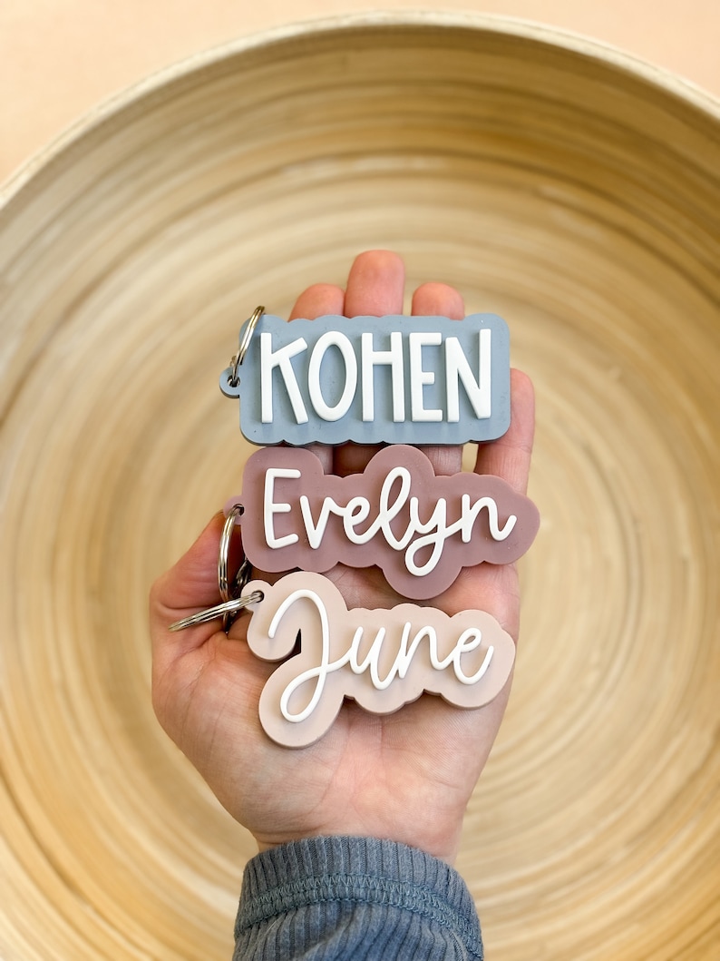 Op de afbeelding: Drie gepersonaliseerde sleutelhangers met de namen "KOHEN", "Evelyn" en "June". "KOHEN" in wit op een blauwgrijze achtergrond. "Evelyn" in witte letters op een mauve achtergrond. "June" in witte letters op een lichtperzik achtergrond.