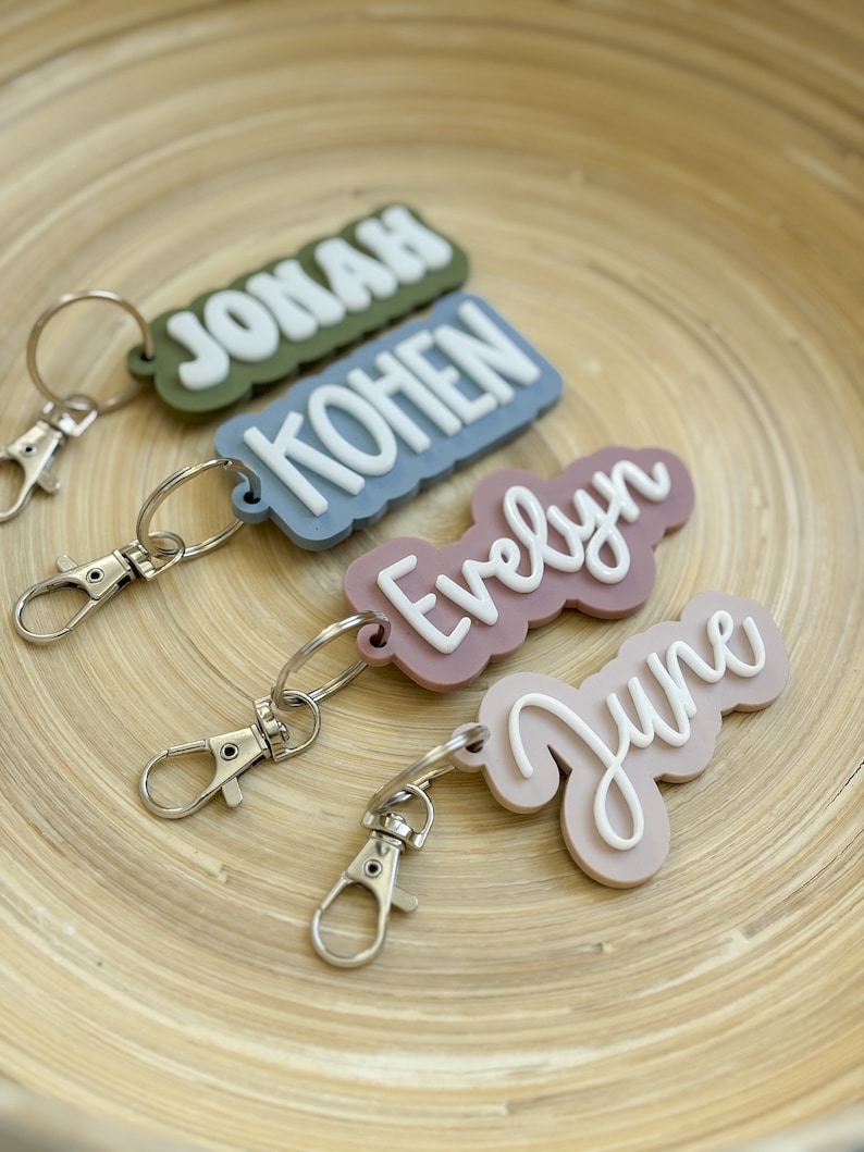 Op de afbeelding: Vier gepersonaliseerde sleutelhangers met de namen: Jonah, Kohen, Evelyn en June. Elke sleutelhanger heeft een andere kleur basis met een contrasterende witte naam. De sleutelhangers hebben zilverkleurige sluitingen.