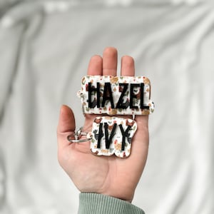 Op de afbeelding: Twee gepersonaliseerde sleutelhangers met de namen "HAZEL" en "IVY". De sleutelhangers hebben een witte achtergrond met een herhaald patroon van kippen in verschillende kleuren. De namen zijn in zwarte letters.