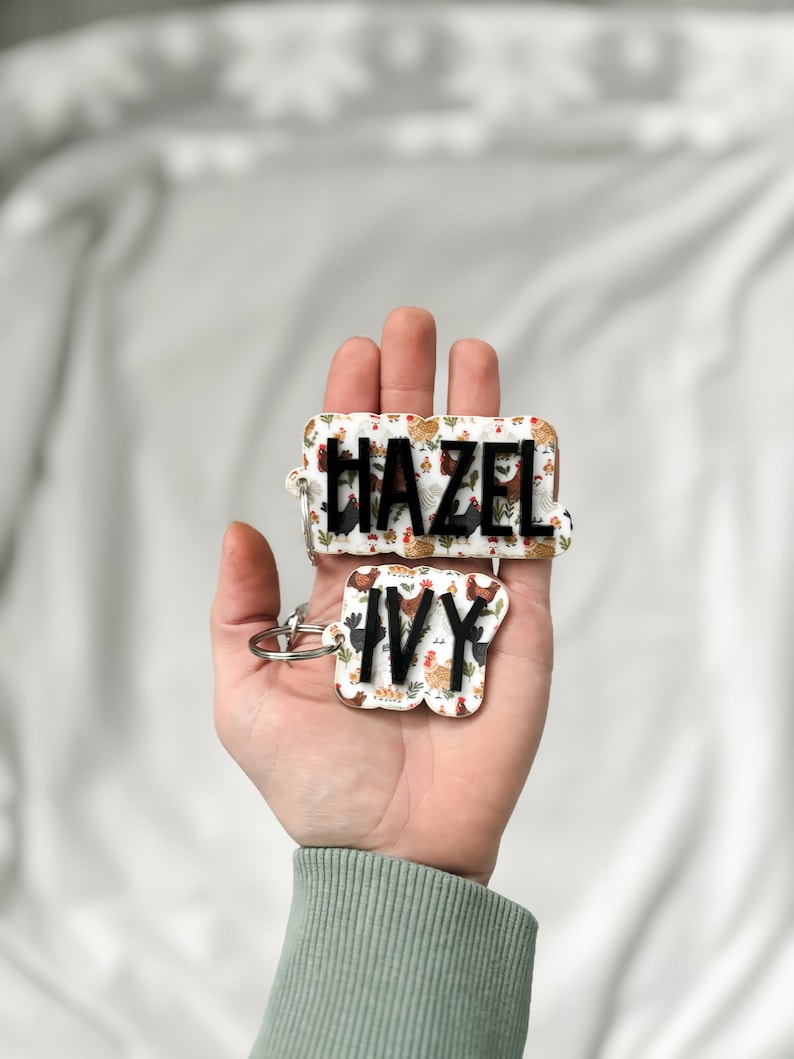 Op de afbeelding: Twee gepersonaliseerde sleutelhangers met de namen "HAZEL" en "IVY". De sleutelhangers hebben een witte achtergrond met een herhaald patroon van kippen in verschillende kleuren. De namen zijn in zwarte letters.
