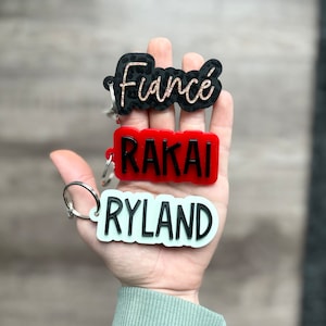 Op de afbeelding: Sleutelhangers met namen en het woord "Fianc&eacute;". De sleutelhangers zijn in zwart, rood en lichtgroen. Het woord "Fianc&eacute;" is in een zwart luipaardprint ontwerp met gouden letters. De andere sleutelhangers hebben de namen "RAKAI" en "RYLAND".